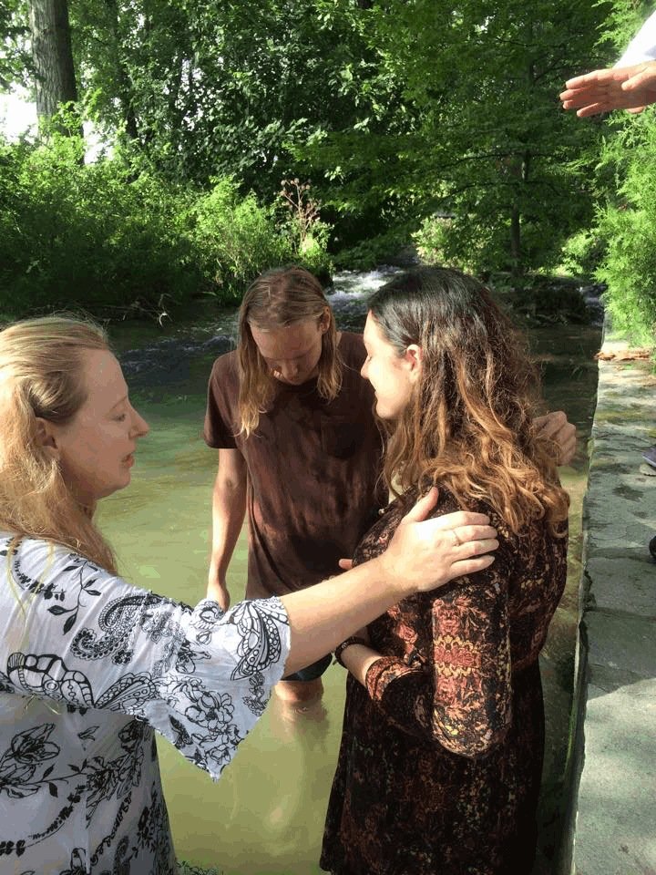 Lyn Baird at a river baptism, standing in the water with a woman and a young person in a pastoral moment of prayer and care.